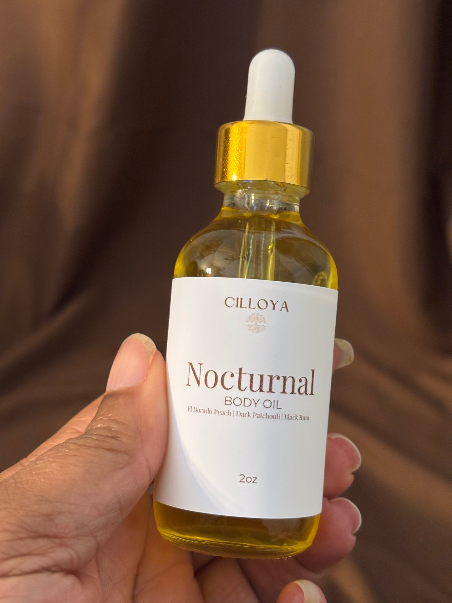 Hand holding a bottle of Cilloya Nocturnal body oil with a dark brown background