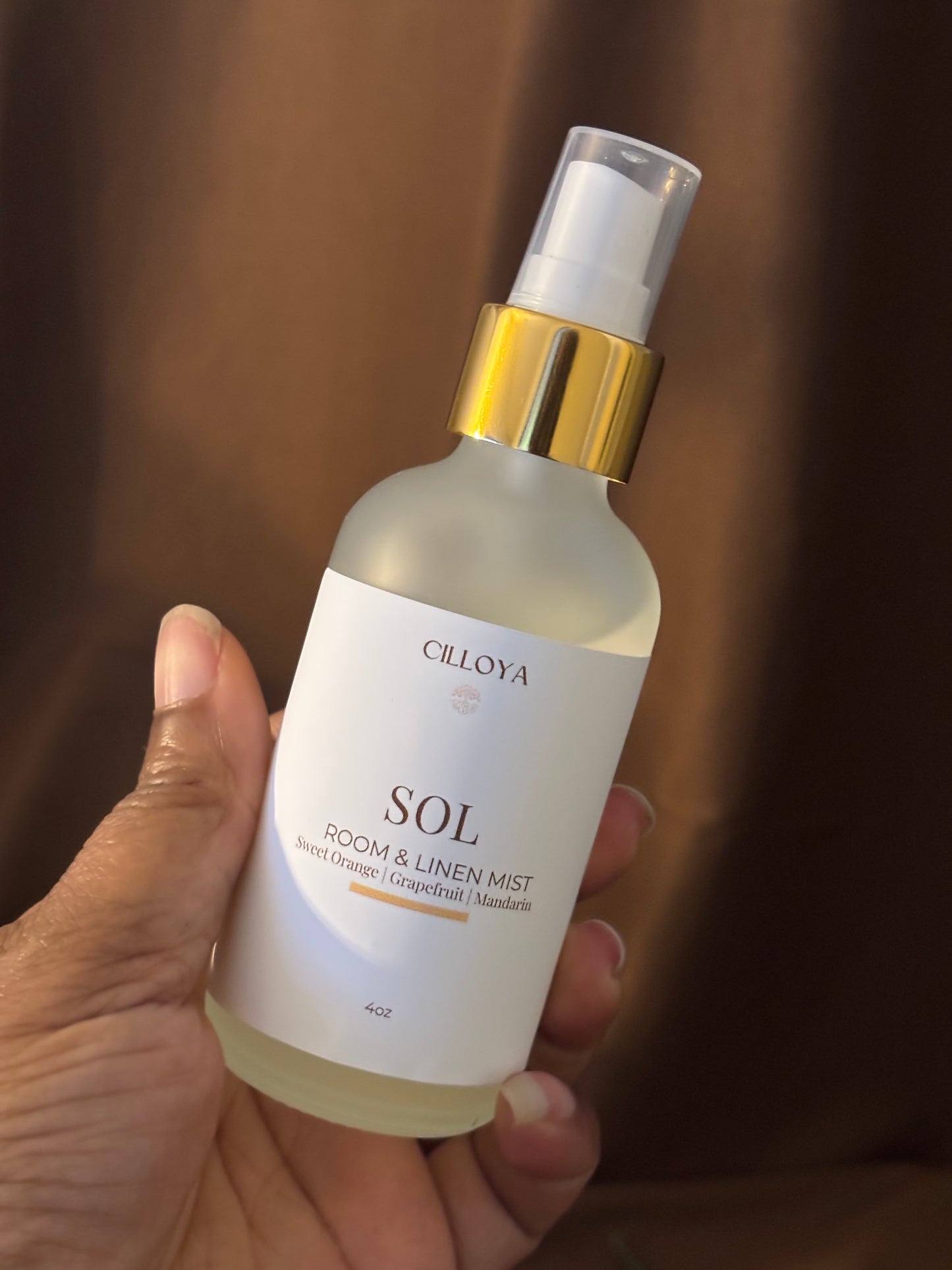 Hand holding a bottle of Cilloya Sol Room & Linen Mist against a dark brown background