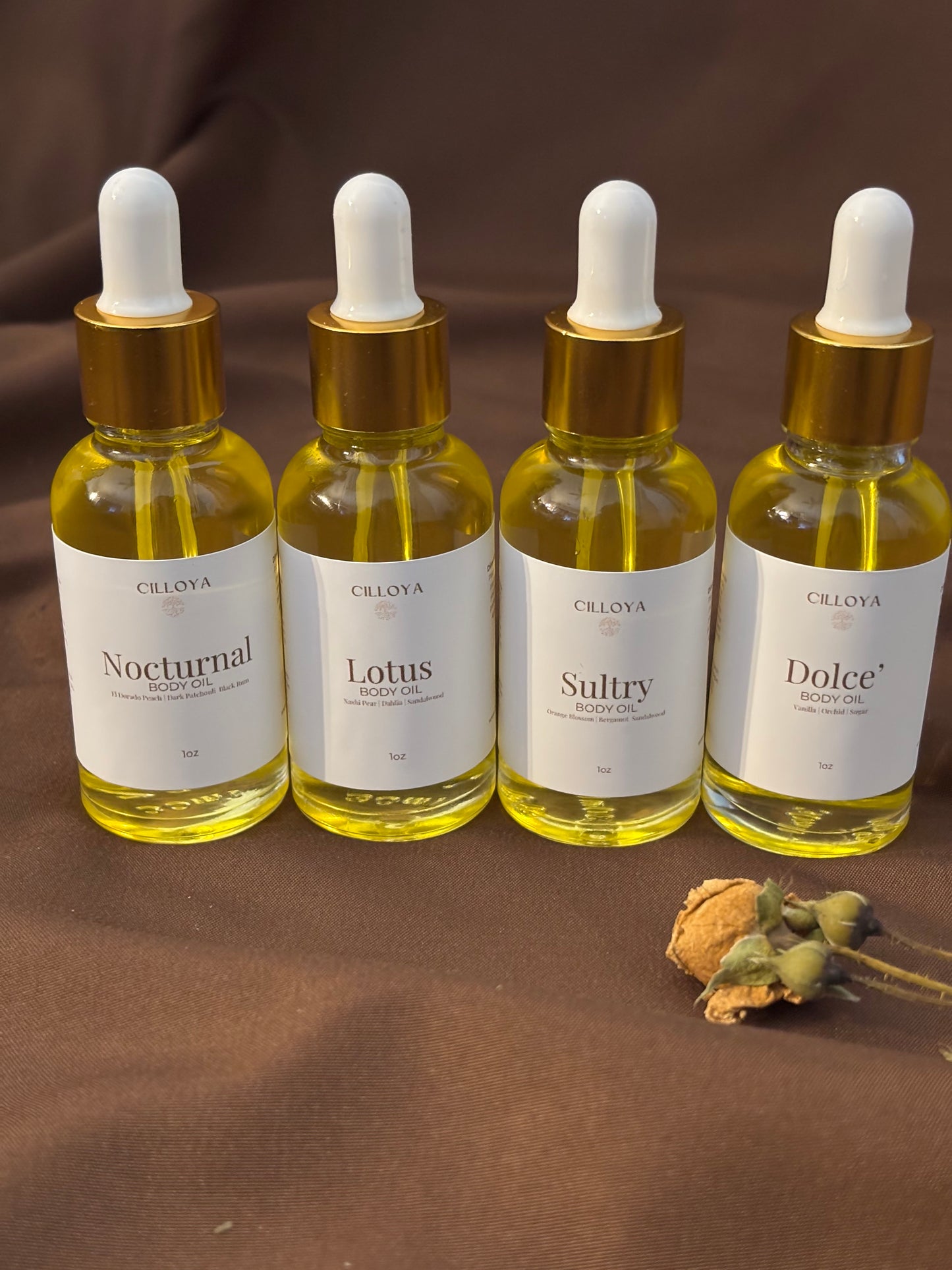 Cilloya Signature Scent Discovery Set featuring four 1oz clear glass dropper bottles with gold necks, including Nocturnal, Lotus, Sultry, and Dolce body oils arranged in a row on a dark brown backdrop.