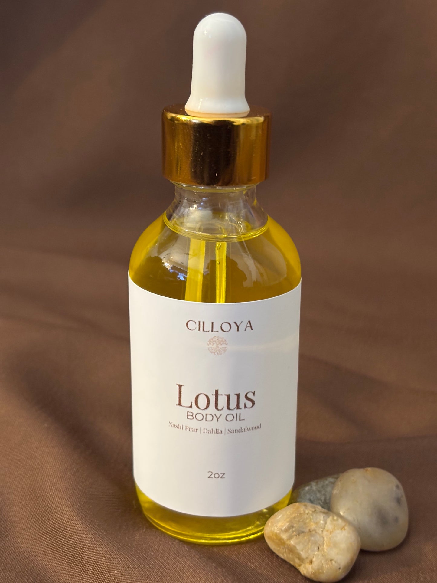 Cilloya Lotus Body Oil in a 2oz clear glass dropper bottle with a gold neck, featuring notes of nashi pear, dahlia and sandalwood on dark brown backdrop