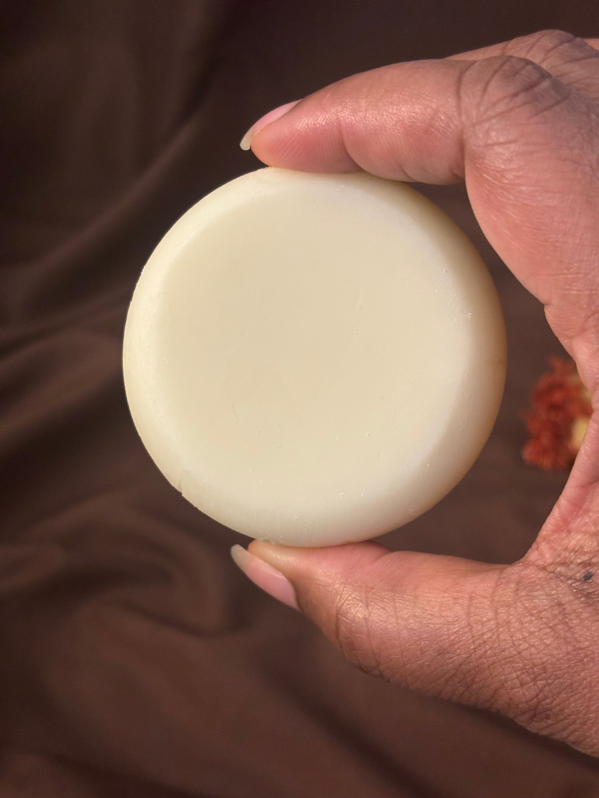 Hand holding  a puck shaped, ivory colored Cilloya Cocoa Butter Bar. against a dark brown backdrop.