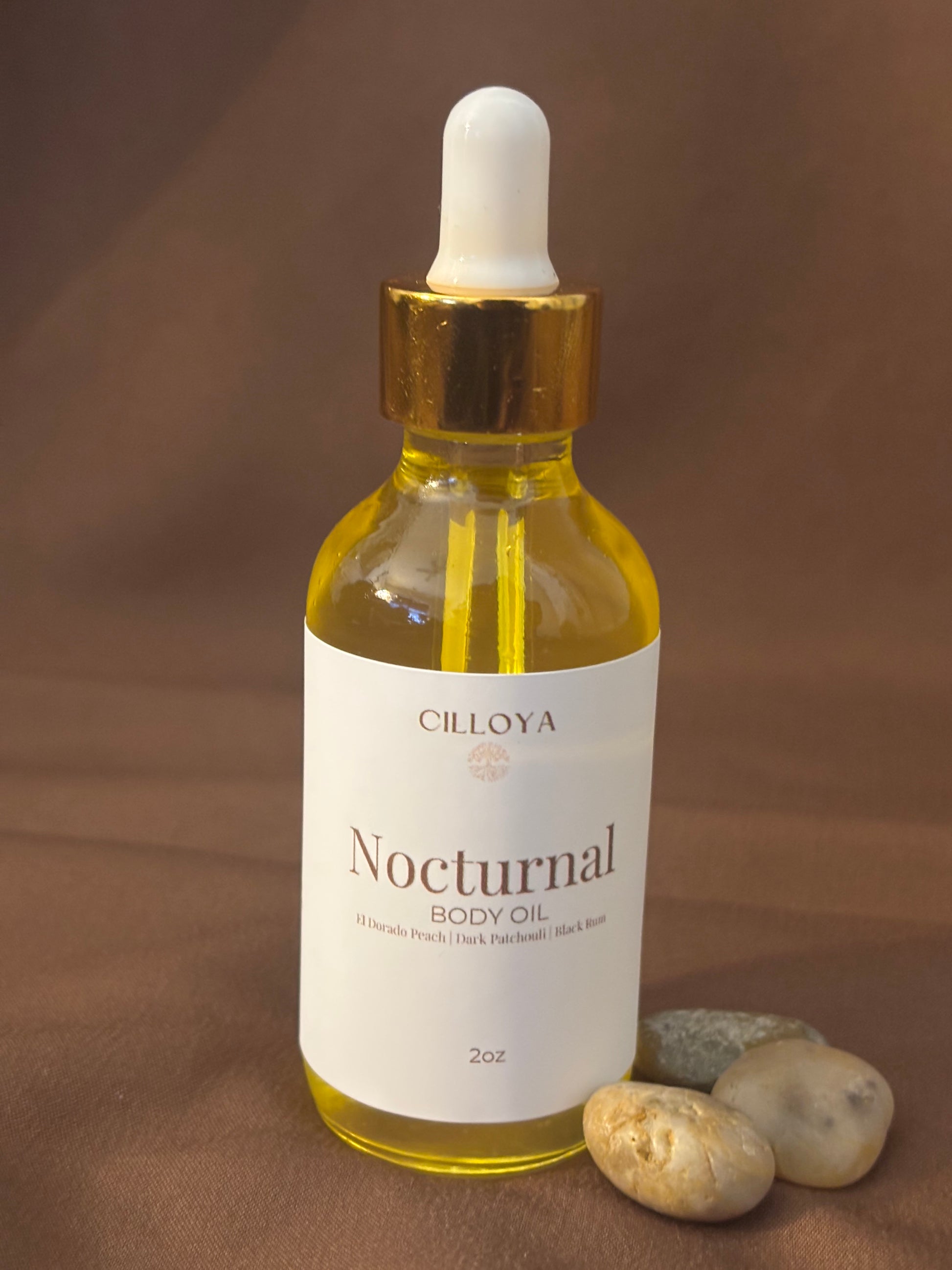 Cilloya Nocturnal Body Oil in a 2oz clear glass dropper bottle with a gold neck, featuring notes of El Dorado Peach, Dark Pathcouli and Black Rum on a dark brown backdrop.