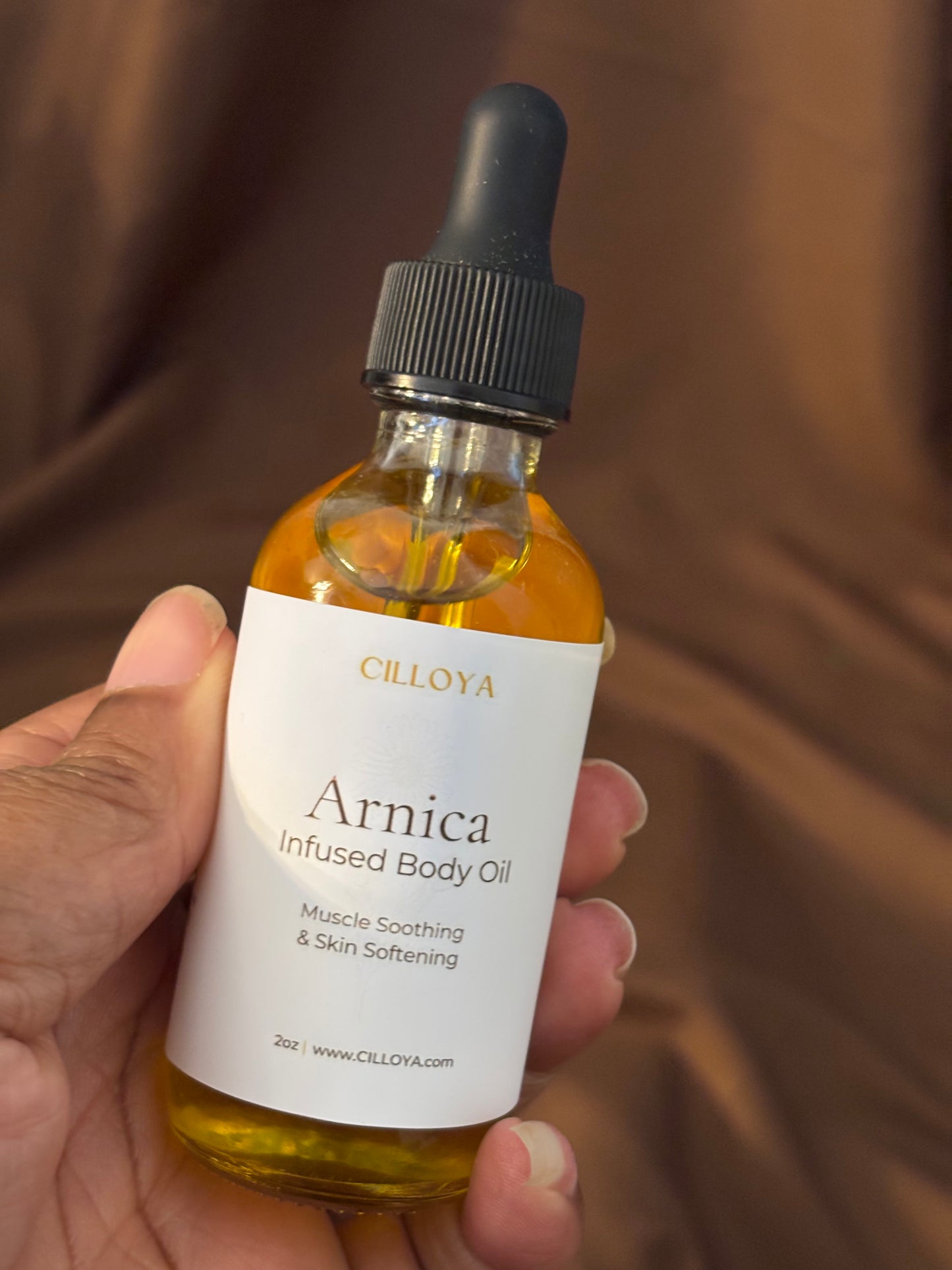 Hand holding a bottle of Cilloya Arnica Infused Body Oil against a brown background