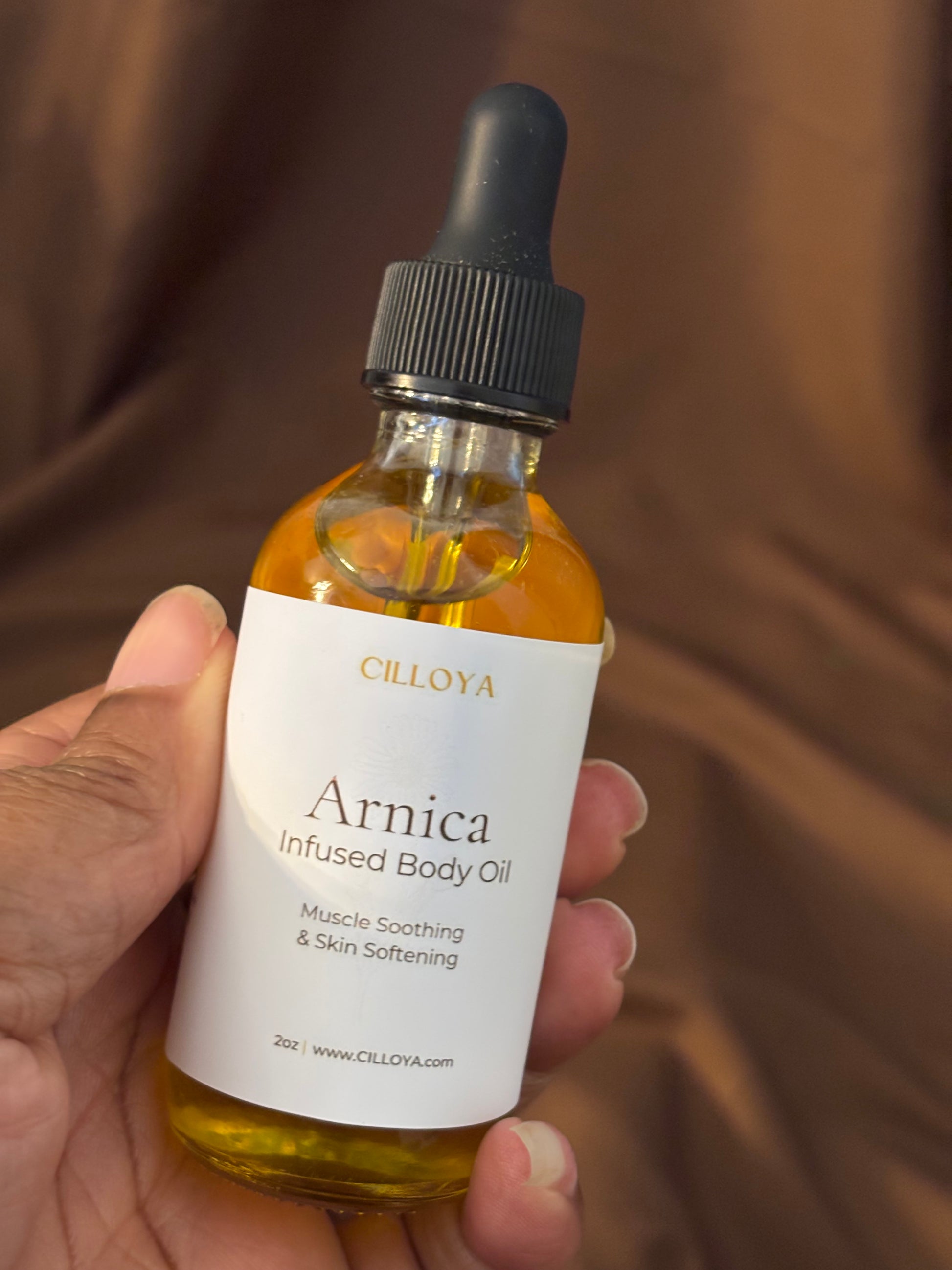Hand holding a bottle of Cilloya Arnica Infused Body Oil against a brown background