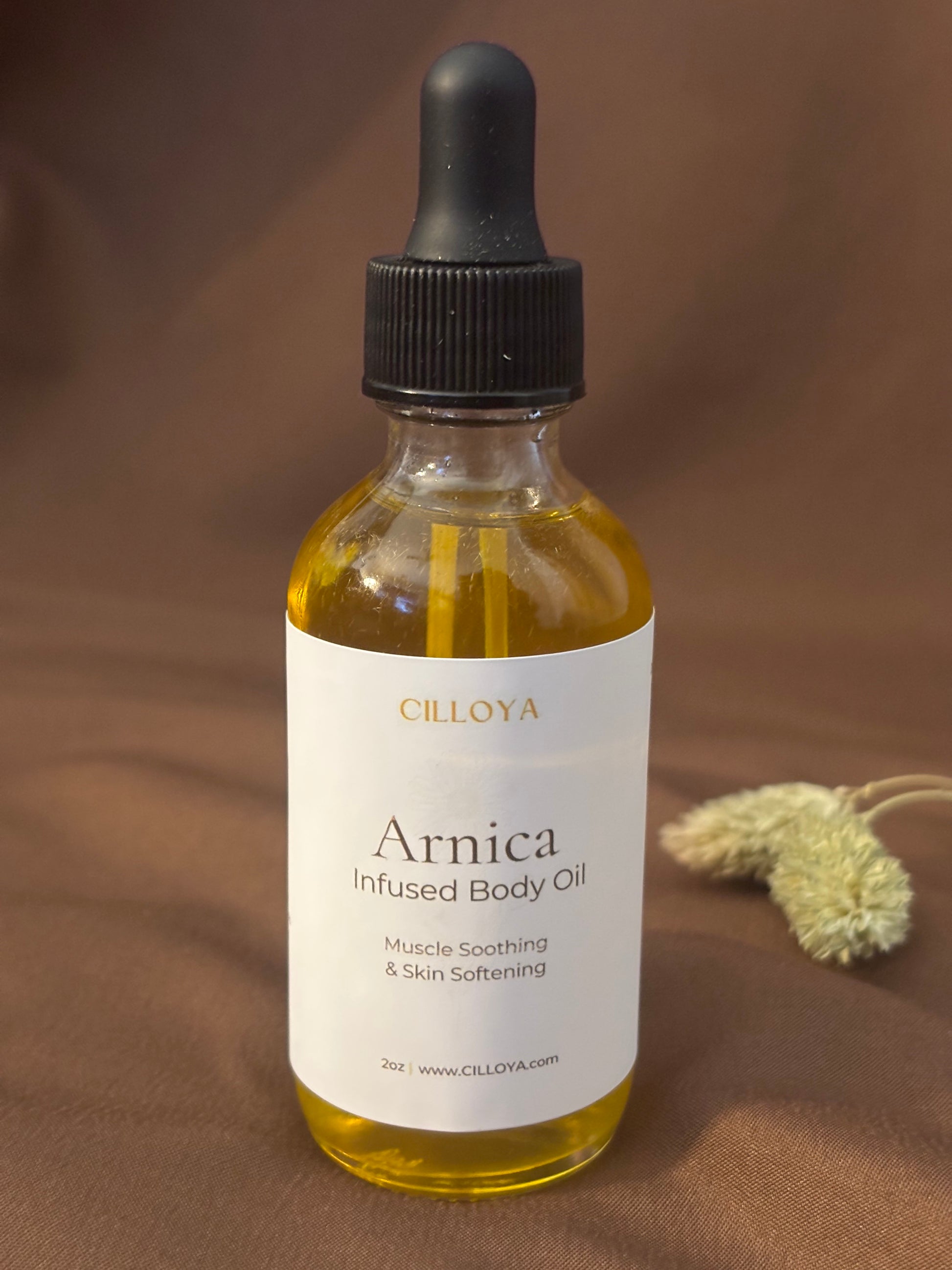 Cilloya Arnica Infused Body Oil in a 2oz clear glass dropper bottle with a black neck on a dark brown backdrop.