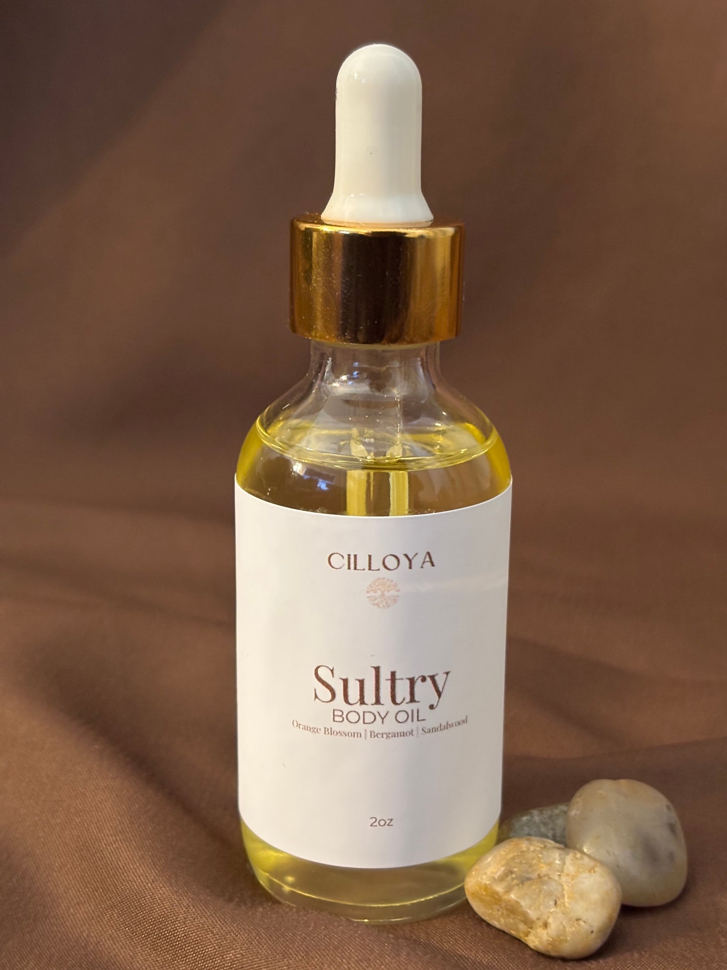 Cilloya Sultry Body Oil in a 2oz clear glass dropper bottle with a gold neck, featuring notes of orange blossom, bergamot and sandalwood on a dark brown backdrop.