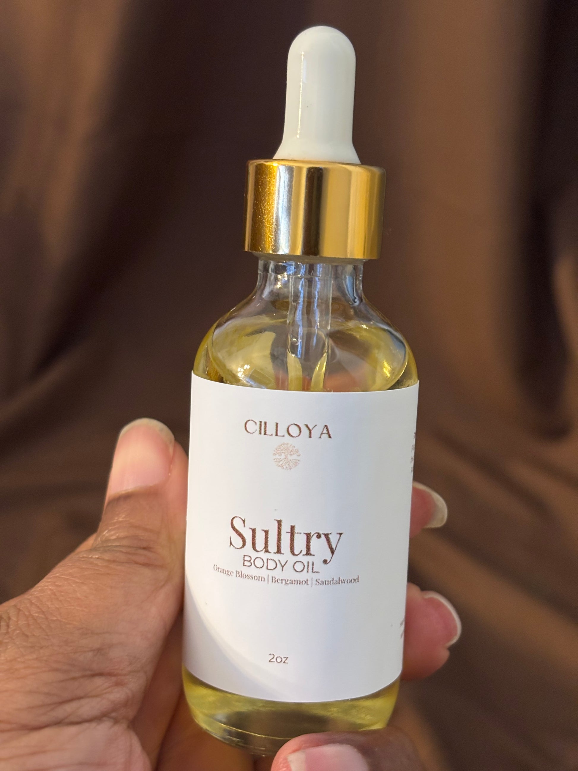Bottle of Cilloya Sultry body oil held by a hand against a brown background